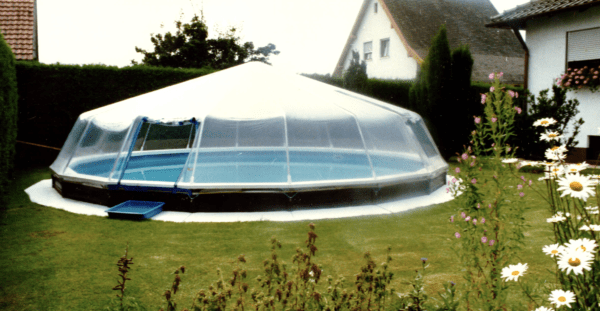 A large transparent Round Above Ground Sun Dome For Steel Wall Pools covering a swimming pool in a backyard, surrounded by a lush garden with blooming flowers.